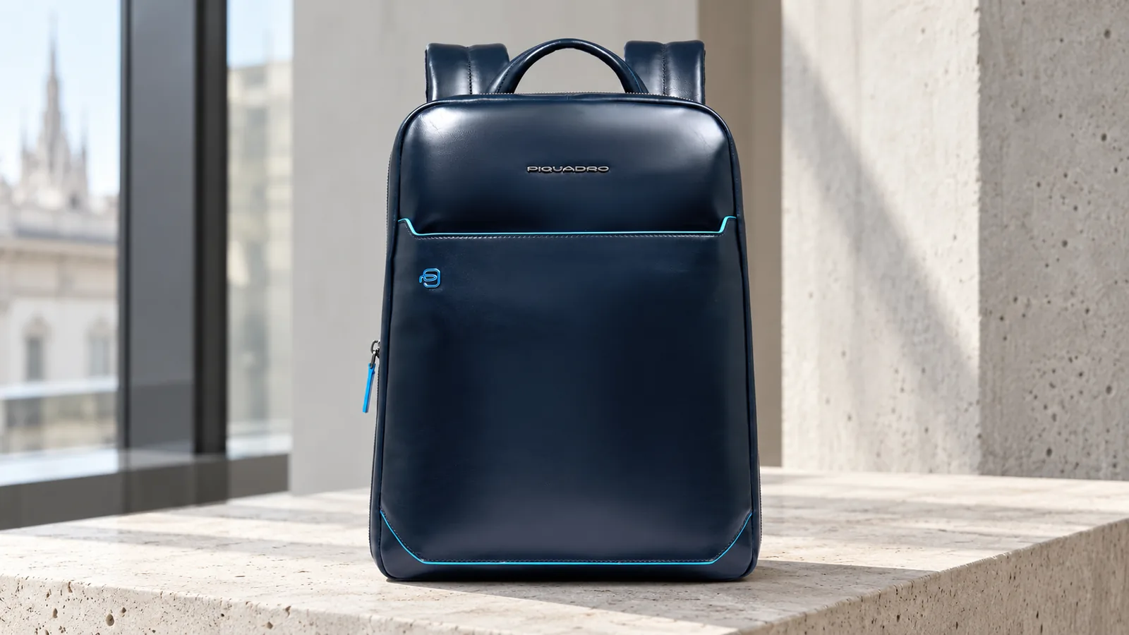 Zaino Piquadro in pelle blu con profili azzurri, accanto a una vetrata.